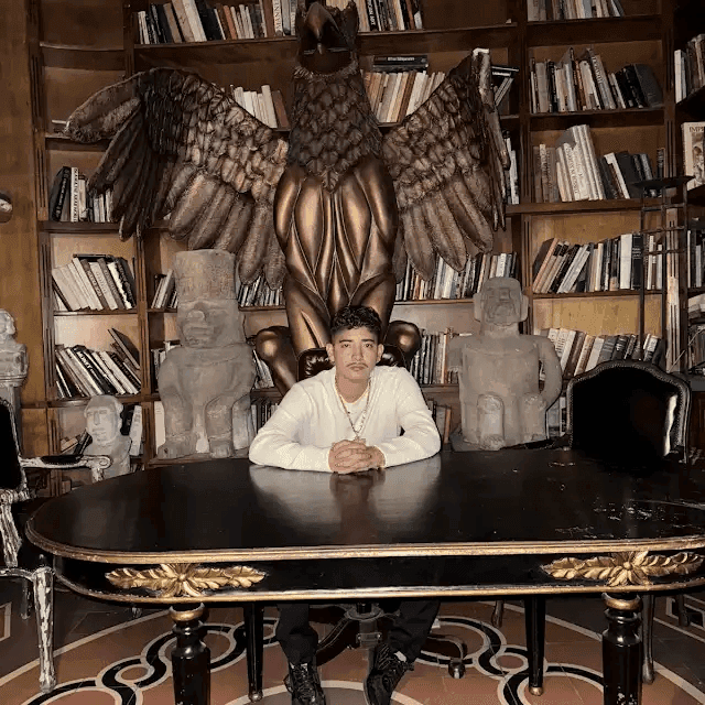 Person sitting at a large wooden desk with eagle statue, surrounded by bookshelves and sculptures.
