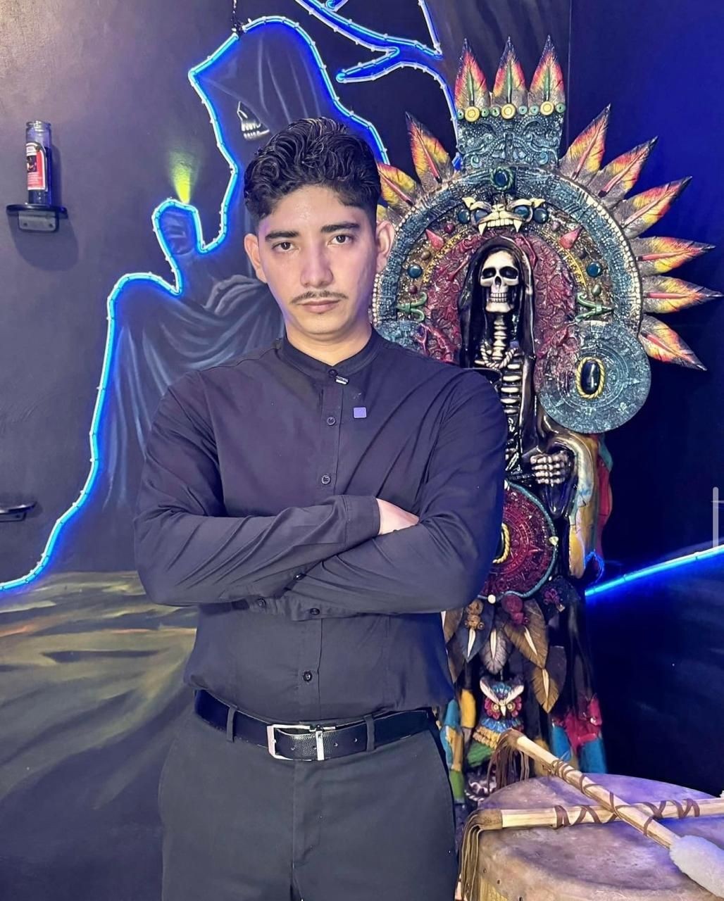 Man in black shirt with arms crossed, neon skeleton mural and colorful skeleton statue in background.