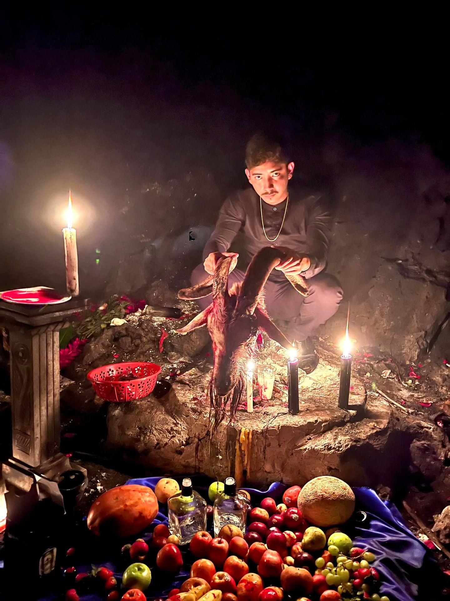 Person performing a ritual with candles, fruit, and animal head in a dimly lit setting.